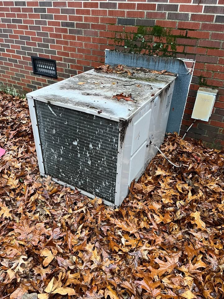 Old, dirty package unit covered in grime and surrounded by leaves beside a brick house